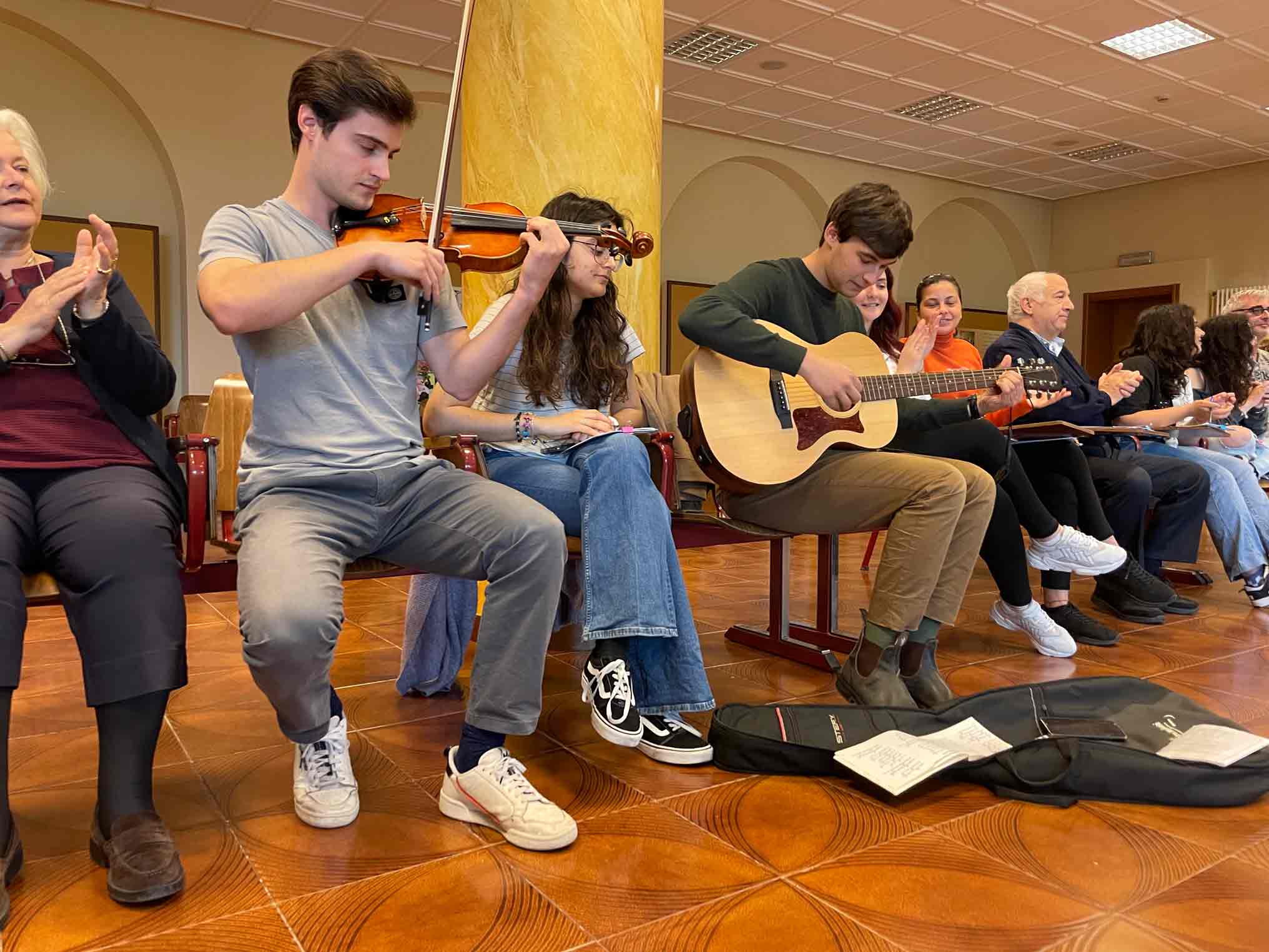 Two young people play violin and guitar next to other seated participants